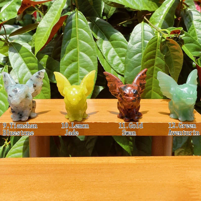 May include: Four small gargoyle figurines carved from different types of stone. The gargoyles are green, yellow, brown, and white with black speckles. The gargoyles are arranged in a row on a wooden surface. The text below each gargoyle identifies the type of stone used to make the figurine.