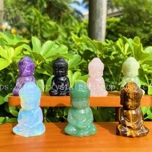 May include: Seven small crystal figurines of Buddha in various colors, including amethyst, black obsidian, rose quartz, green aventurine, opalite, and tiger's eye. The figurines are arranged on a wooden shelf.
