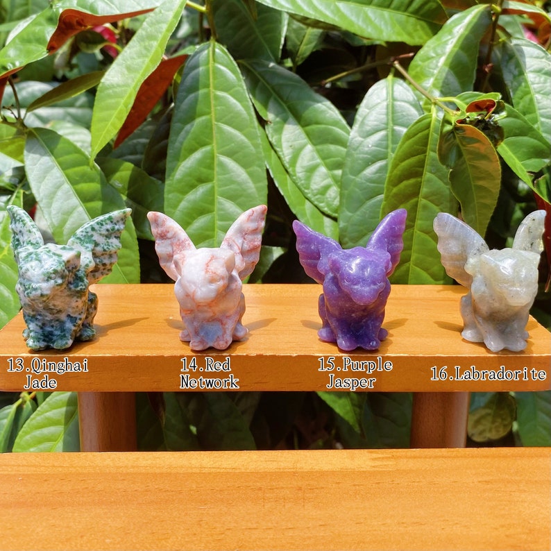 May include: Four small carved stone gargoyle figurines, each with wings and a different colour: green, pink, purple, and grey. The gargoyles are sitting on a wooden shelf with labels underneath: 13. Qinghai Jade, 14. Red Network, 15. Purple Jasper, 16. Labradorite.