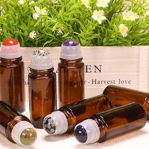 Pode incluir: Uma coleção de frascos de vidro âmbar com esferas de pedras preciosas em cores que incluem vermelho, branco com manchas pretas, roxo, verde e azul escuro com brilhos, cada um com uma tampa de plástico transparente sobre a esfera.