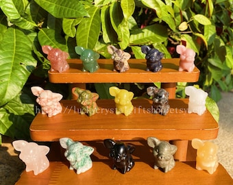 May include: A collection of small, carved crystal elephant figurines in various colors, including pink, green, yellow, and black. The elephants are displayed on wooden shelves against a backdrop of green foliage.