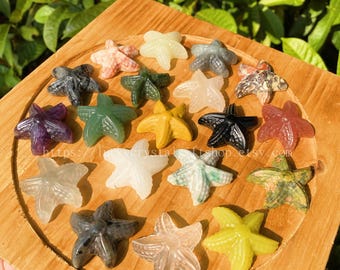 May include: A collection of carved starfish figurines in various colors, including green, yellow, black, and clear. The starfish are arranged on a wooden tray, with a natural green background. The Etsy shop URL is visible on the tray.