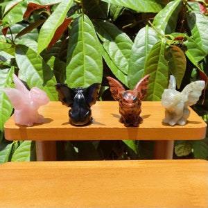 May include: Four small carved gargoyle figurines made of different types of stone. The gargoyles are pink, black, red, and grey. They are sitting on a wooden shelf.