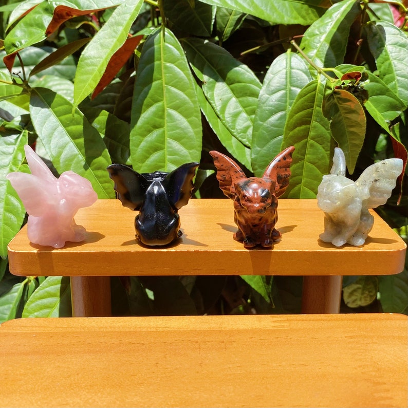 May include: Four small carved gargoyle figurines made of different types of stone. The gargoyles are pink, black, red, and grey. They are sitting on a wooden shelf.