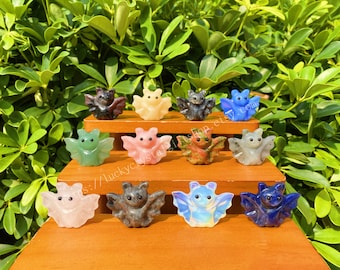 May include: A collection of 12 hand-carved crystal bat figurines in various colors, including pink, green, blue, and black. The bats are arranged on a wooden shelf, with each bat facing forward.