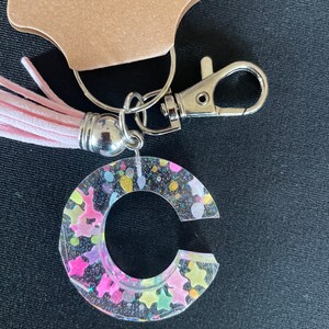 May include: A clear keychain with a pink tassel and a silver charm. The keychain is shaped like the letter C and is filled with colorful glitter and stars.