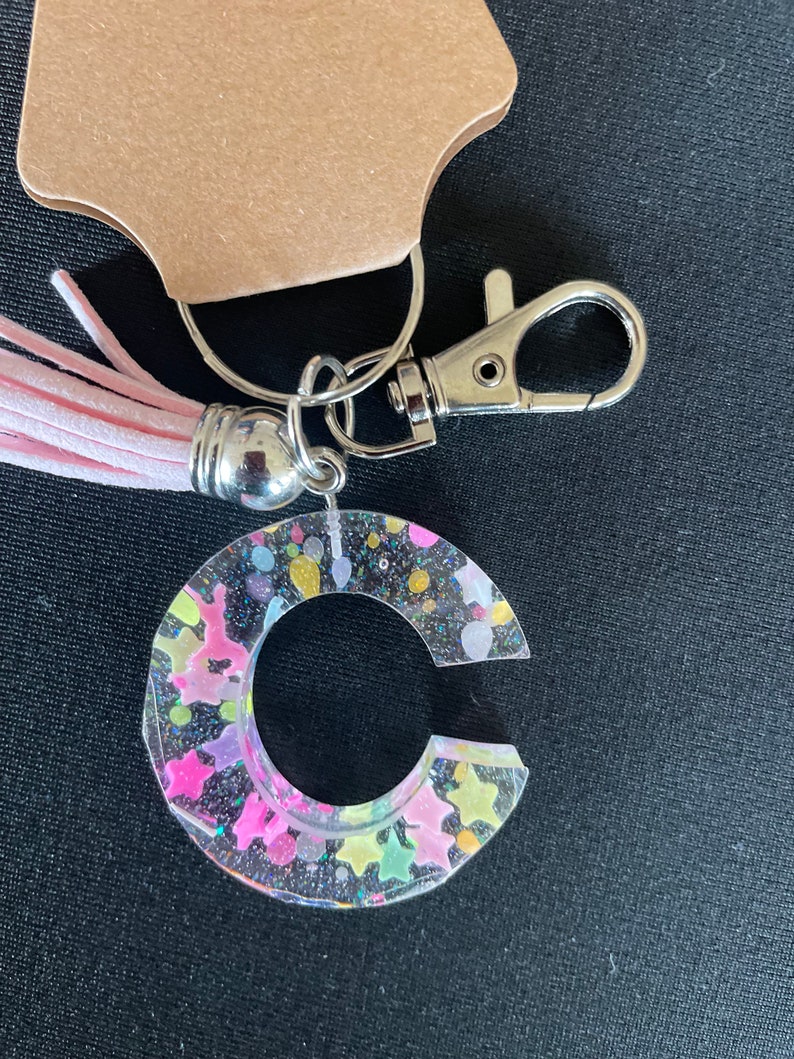 May include: A clear keychain with a pink tassel and a silver charm. The keychain is shaped like the letter C and is filled with colorful glitter and stars.