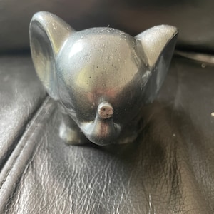 Puede incluir: Una pequeña figurita de elefante negro con un acabado liso y brillante. La trompa del elefante está enroscada y sus orejas son grandes y caídas.