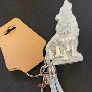 Può includere: Un portachiavi a forma di lupo color argento con un disegno di lupo che ulula, alberi e stelle. Il portachiavi ha un fermaglio argentato, un nappa argentata e un'etichetta marrone.