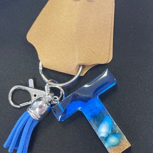 May include: Blue resin letter T keychain with a blue tassel and seashells embedded in the resin. The keychain is attached to a silver ring and a silver lobster clasp.