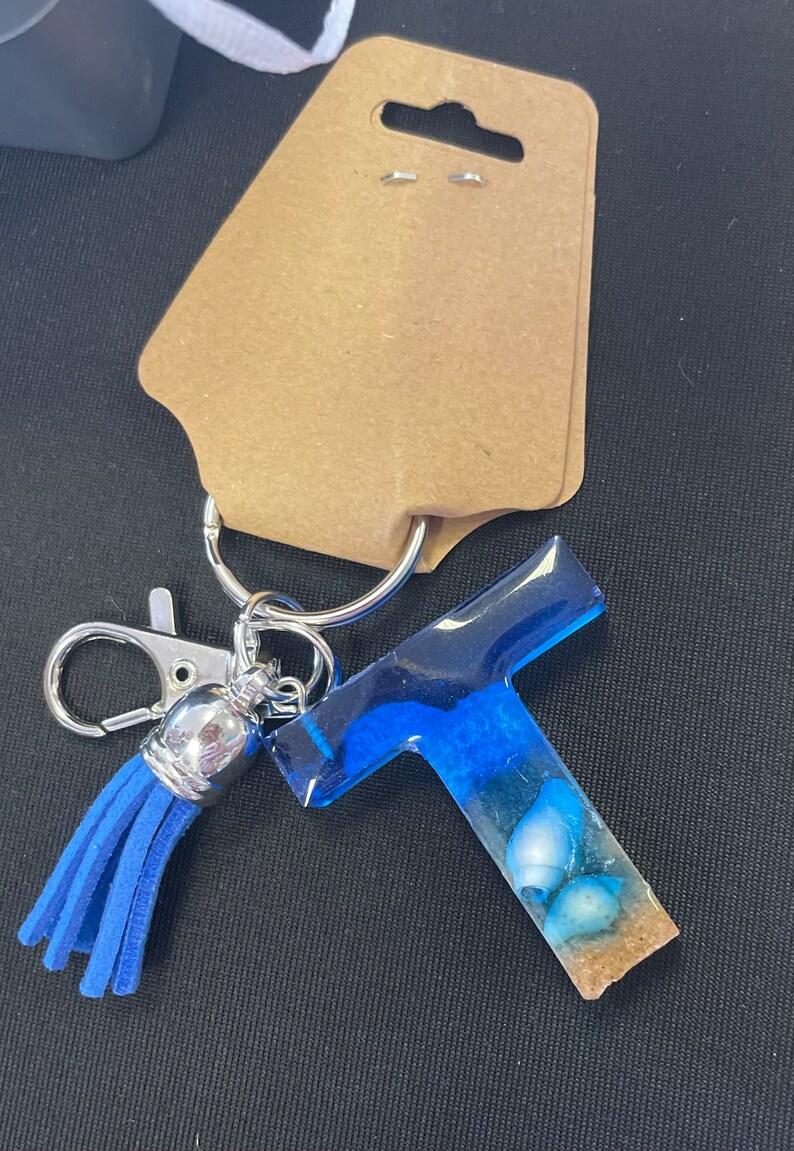 May include: Blue resin letter T keychain with a blue tassel and seashells embedded in the resin. The keychain is attached to a silver ring and a silver lobster clasp.