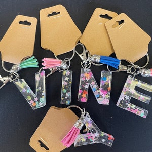 May include: Keychains with the letters VINEA, each letter is filled with colorful glitter and has a small tassel attached. The keychains are on brown card stock.