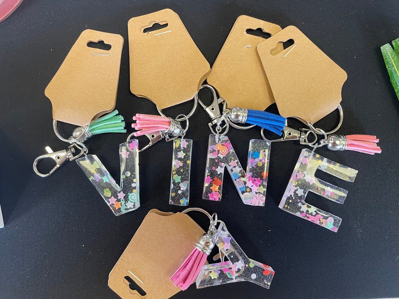 May include: Keychains with the letters VINEA, each letter is filled with colorful glitter and has a small tassel attached. The keychains are on brown card stock.
