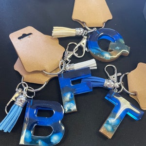 May include: Five resin letter keychains with blue and white seashell designs. Each keychain has a silver metal ring and a tassel. The letters are B, F, Q, and T.