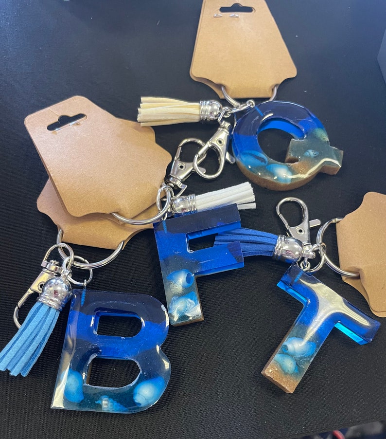 May include: Five resin letter keychains with blue and white seashell designs. Each keychain has a silver metal ring and a tassel. The letters are B, F, Q, and T.