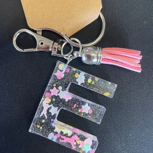 May include: A clear resin keychain with the letter "E" embedded in it. The letter is filled with colorful glitter, stars, and confetti. A pink tassel hangs from the keychain.
