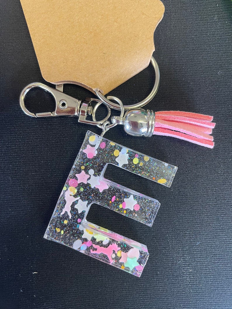 May include: A clear resin keychain with the letter "E" embedded in it. The letter is filled with colorful glitter, stars, and confetti. A pink tassel hangs from the keychain.