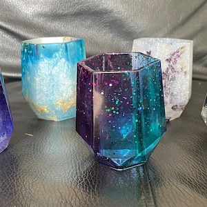 Puede incluir: Colección de cinco vasos decorativos de resina. Cada vaso tiene una combinación de colores única, incluyendo tonos de azul, morado y turquesa, con detalles de purpurina. Los vasos tienen un diseño facetado y son adecuados para diversos usos.