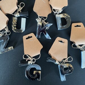 May include: A set of ten resin letter keychains with black and white marbled designs. Each keychain features a different letter of the alphabet and a silver key ring with a tassel.