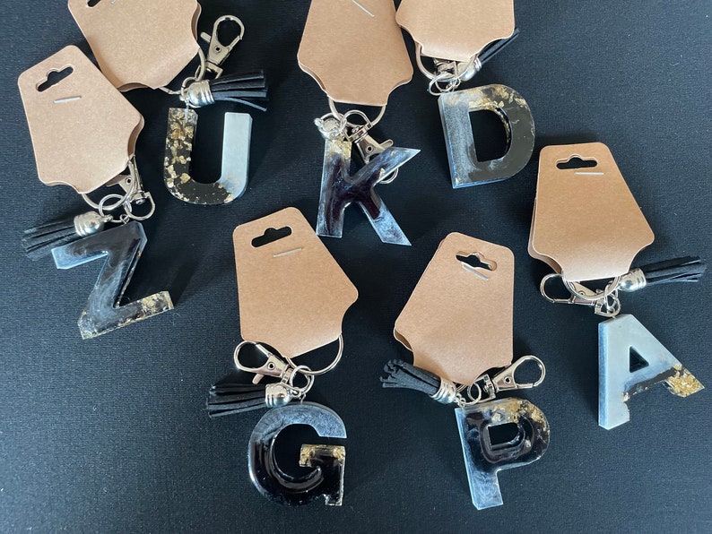 May include: A set of ten resin letter keychains with black and white marbled designs. Each keychain features a different letter of the alphabet and a silver key ring with a tassel.