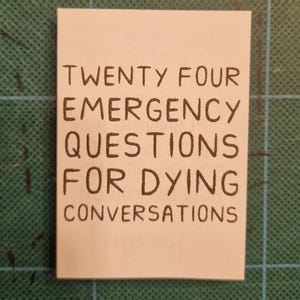 May include: A small, rectangular white card with black handwritten text. The text reads "TWENTY FOUR EMERGENCY QUESTIONS FOR DYING CONVERSATIONS". The card is on a green cutting mat.
