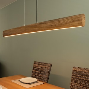 Può includere: Lampada a sospensione rettangolare in legno. La luce è una lunga trave orizzontale in legno naturale con una striscia di luce calda e brillante nella parte inferiore. Lo sfondo è verde tenue. La lampada è lunga circa 120 cm.