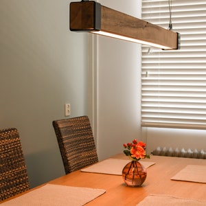 Op de afbeelding: Een moderne houten balk hanglamp met zwarte metalen accenten, boven een eettafel. De tafel is gedekt met placemats, stoelen en een vaas met bloemen. De kamer heeft een neutraal kleurenpalet.