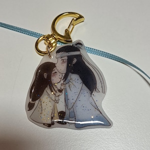 Può includere: Un portachiavi trasparente con un moschettone dorato che presenta un'immagine di cartone animato di due persone in abiti tradizionali cinesi. L'immagine ha dettagli glitter.