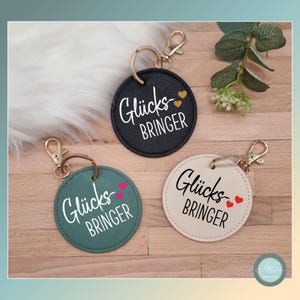 Könnte beinhalten: Drei runde Leder-Schlüsselanhänger mit goldenen Ringen. Jeder Schlüsselanhänger hat den Text "Glücksbringer" mit einem Herz-Design. Die Schlüsselanhänger sind schwarz, grün und beige.