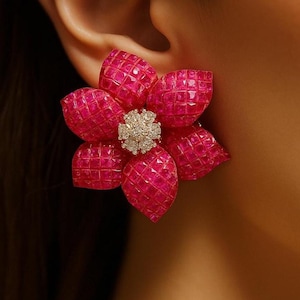 Puede incluir: Primer plano de un pendiente con forma de flor. El pendiente presenta cinco pétalos de color rosa brillante con una superficie texturizada. El centro de la flor es un grupo de pequeñas piedras brillantes. El pendiente está sobre un fondo borroso.