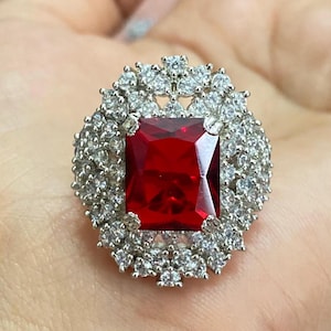 Peut inclure: Une bague argentée ornée d'une grande pierre rouge vif de forme carrée en son centre. La pierre rouge est entourée d'un halo de petites pierres rondes et transparentes, créant un design étincelant. La bague est une pièce maîtresse.