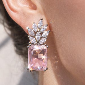 Può includere: Primo piano di un orecchino scintillante. L'orecchino presenta una grande gemma rosa rettangolare alla base, sormontata da un gruppo di pietre chiare a taglio marquise. L'incastonatura è color argento e l'orecchino è indossato su un orecchio.