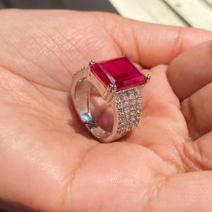 以下が含まれることがあります： 大きな長方形のルビーレッドの宝石をあしらったシルバーリング。バンドには小さな透明な宝石が飾られています。リングは手で持たれており、そのデザインと色を際立たせています。