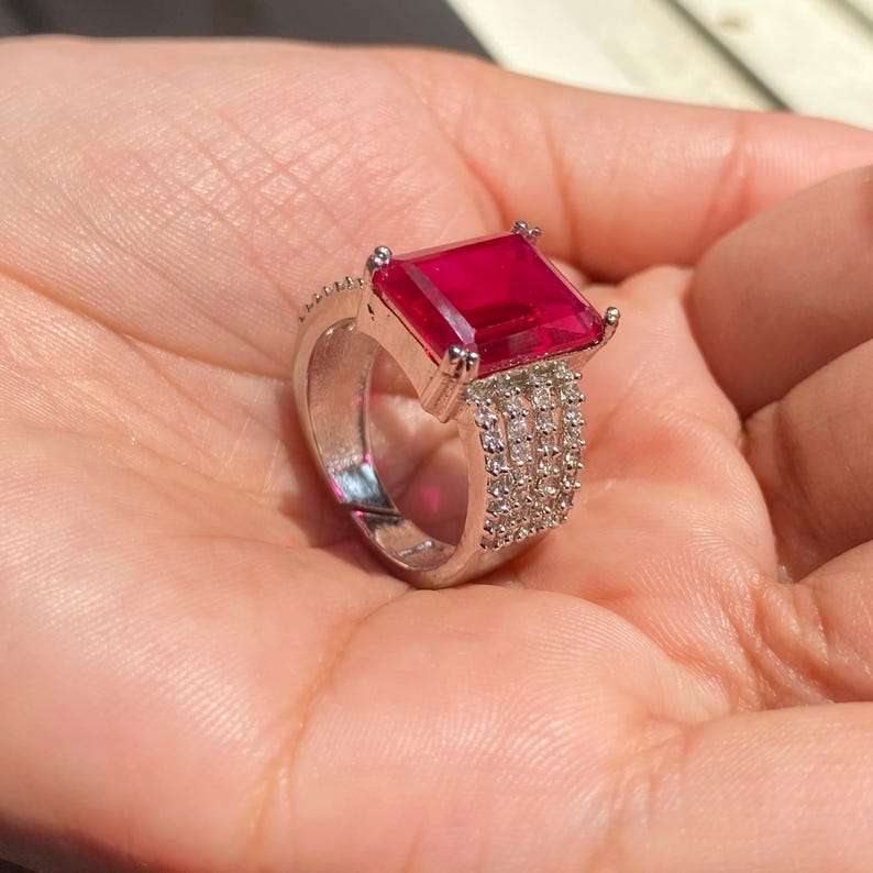 以下が含まれることがあります： 大きな長方形のルビーレッドの宝石をあしらったシルバーリング。バンドには小さな透明な宝石が飾られています。リングは手で持たれており、そのデザインと色を際立たせています。