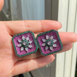 May include: A pair of square stud earrings with a floral design. The earrings feature a magenta center surrounded by green and white gemstones. The outer edge is green, and the center has a white flower design.