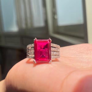 以下が含まれることがあります： 大きな長方形の鮮やかなピンク色の宝石をあしらったシルバーリング。バンドには、小さな透明な宝石が複数列並んでいます。このリングは、大胆で鮮やかなデザインを特徴とする、ステートメントピースです。