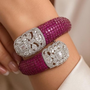 Può includere: Due braccialetti di lusso. I braccialetti presentano una fascia strutturata color magenta intenso e un elemento centrale argentato tempestato di diamanti. I diamanti sono incastonati in un intricato motivo floreale. I braccialetti sono indossati al polso.