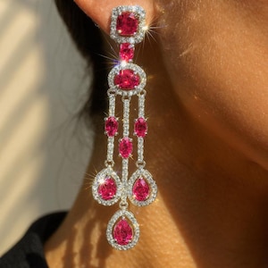May include: Elegant drop earrings with a square top, oval centre, and teardrop bottom. The earrings feature bright pink gemstones surrounded by a halo of small, clear stones. The setting is silver-toned, and the design is intricate.