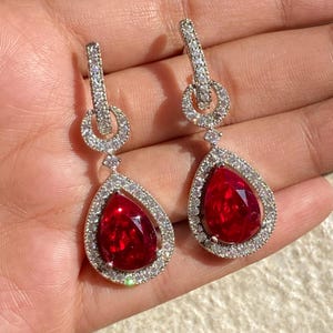Puede incluir: Un par de pendientes de plata con una piedra preciosa roja en forma de lágrima rodeada por un halo de piedras preciosas transparentes más pequeñas.