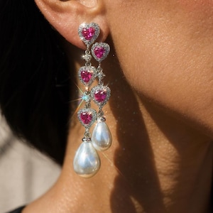 Puede incluir: Pendientes colgantes con gemas rosas en forma de corazón, rodeadas de pequeñas piedras transparentes. Los pendientes tienen un engaste de metal plateado y terminan con una gran perla blanca en forma de pera.