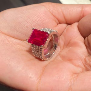 以下が含まれることがあります： 大きな正方形のルビーレッドの宝石が付いたシルバーリング。バンドは小さな透明な石で装飾されています。リングは手で持たれており、ジュエリーのデザインと色を際立たせています。