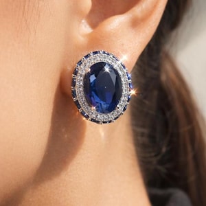 Può includere: Orecchino a lobo di forma ovale con una grande gemma blu scuro al centro, circondata da un alone di gemme più piccole e trasparenti. L'orecchino è incastonato in un metallo color argento, creando un aspetto scintillante ed elegante.