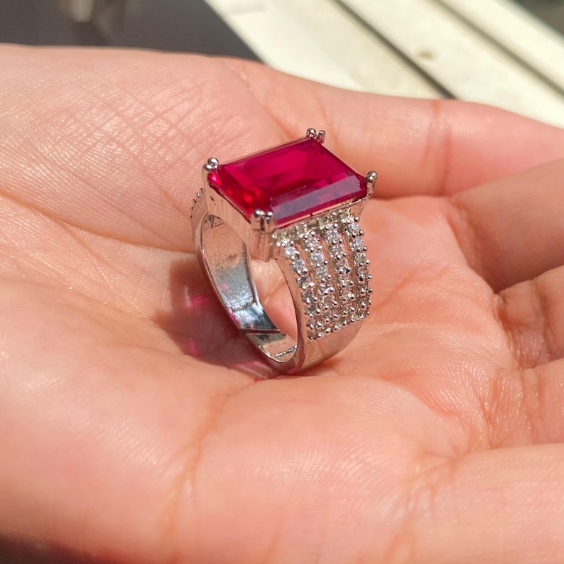 以下が含まれることがあります： 大きな長方形の鮮やかな赤い宝石をあしらったシルバーリング。リングのバンドには、小さな透明な石が複数列並んでいます。リングは開いた手のひらに飾られ、そのデザインと輝きを際立たせています。