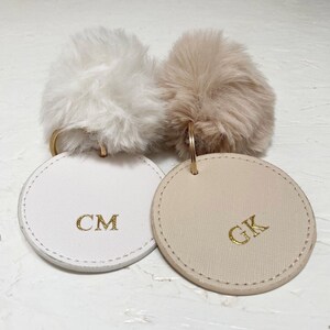 May include: Two round leather keychains with fluffy pom poms. The keychain on the left is white with the initials "CM" in gold lettering. The keychain on the right is beige with the initials "GK" in gold lettering.