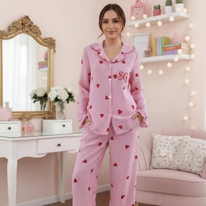 May include: Pink striped pajama set with red heart accents. The long-sleeved top has a ruffled collar and the initials "SG" embroidered on the chest. The matching pants are wide-legged. The set is made of a soft, comfortable material.