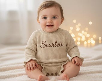 Könnte beinhalten: Ein Baby in einem beigefarbenen Strickpullover und passenden Shorts, beide mit Rüschenkanten. Der Name "Scarlett" ist auf den Pullover gestickt. Das Baby sitzt auf einer weichen, weißen Decke und lächelt in die Kamera.