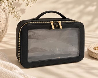 Puede incluir: Un estuche de viaje rectangular negro con un panel frontal transparente y un asa superior. El estuche cuenta con un cierre de cremallera doble en tono dorado con las iniciales SB grabadas en el tirador. Diseño con estructura rígida y herrajes.