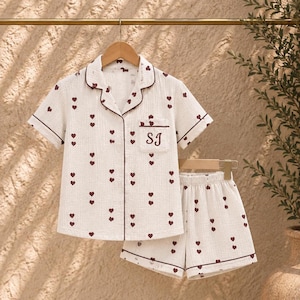 May include: A short-sleeved button-down pajama shirt and matching shorts set in off-white fabric with a small dark red heart pattern. The shirt features a notched collar, dark red piping, and a chest pocket embroidered with the letters SJ.