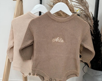 Könnte beinhalten: Zwei gestrickte Baby-Strampler hängen an einem Holzständer. Einer ist hellbeige, der andere braun mit dem Namen "Arlo" auf der Brust gestickt. Beide haben lange Ärmel und Knöpfe. Grobstrick.
