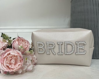 May include: A cream-colored cosmetic bag with the word "BRIDE" spelled out in pearl-like beads. The bag has a gold zipper pull and is next to a bouquet of pink flowers. The bag is a rectangular shape.